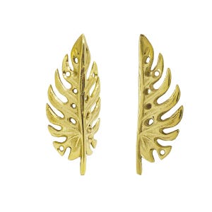 May include: Two gold-colored metal cabinet pulls shaped like monstera leaves. The left pull is a full leaf design, while the right pull is a half-leaf design. Both pieces feature a polished finish with visible screw holes for mounting hardware.