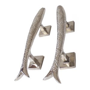 Op de afbeelding: Twee zilverkleurige metalen kastgrepen in de vorm van vissen. Elke handgreep heeft een gestructureerd schubpatroon langs het lichaam, een gebogen silhouet en twee vierkante montagevoeten die aan de achterkant uitsteken voor bevestiging.