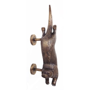 Puede incluir: Un tirador de puerta de metal en tono bronce con forma de nutria vertical. La nutria se muestra en posición de nado con el cuerpo alargado, la cola hacia arriba y las cuatro patas extendidas. Se fija a la puerta mediante dos placas circulares.