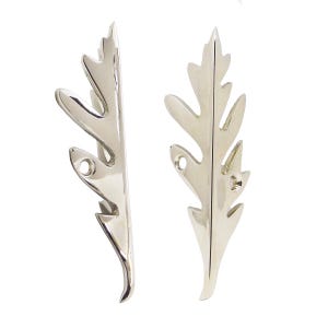 May include: Two polished silver-toned metal cabinet handles shaped like elongated, serrated leaves. Each handle features a central spine with pointed, jagged edges and two small circular screw holes for mounting, displaying a smooth, reflective finish.