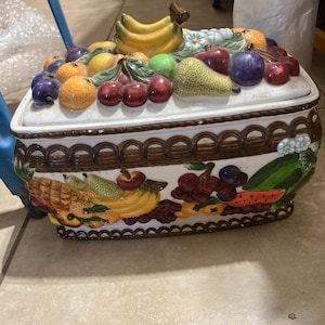 May include: A rectangular ceramic lidded container featuring a three-dimensional fruit motif. The lid is adorned with sculpted bananas, pears, cherries, and oranges. The base displays a brown lattice border and painted depictions of various fruits.