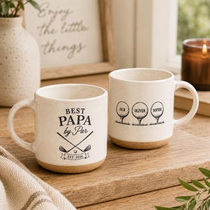 May include: Two off-white ceramic mugs with a speckled finish and a tan base. One mug features the black text "BEST PAPA by Par" with crossed golf clubs and "EST. 2026". The other mug displays three golf balls on tees labeled "JACK", "OLIVER", and "SOPHIE".