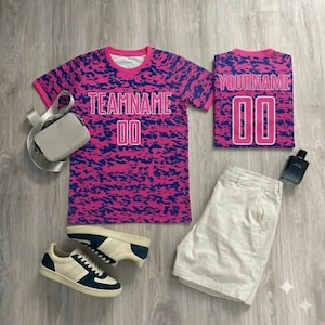 May include: A short-sleeved t-shirt and a folded t-shirt, both featuring a pink and blue camouflage pattern with the text TEAMNAME 00 and YOURNAME 00. Also included are beige shorts, a light gray crossbody bag, and navy and white sneakers.