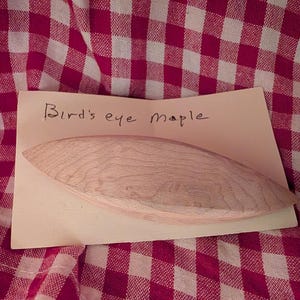 Könnte beinhalten: Ein glattes, helles Holzschiffchen mit einer spitzen, länglich ovalen Form. Das Objekt liegt auf einer rechteckigen Karte, auf der der handschriftliche Text Bird's eye Maple über dem Holzstück zu sehen ist, das eine natürliche Maserung zeigt.