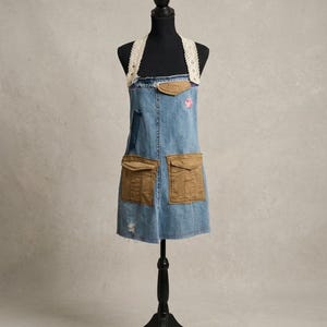 Peut inclure: Un tablier en jean bleu doté de deux grandes poches cargo marron clair sur le devant, d'un petit empiècement marron clair à l'encolure et de larges bretelles en dentelle blanche. Le tissu présente une légère usure et un motif floral rose.