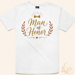 May include: A white short-sleeved t-shirt featuring a gold graphic print on the chest. The design includes a small bow tie icon above the text "Man of Honor" in a cursive script, flanked by two laurel leaf branches and decorative flourishes.