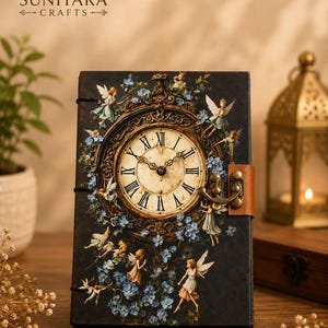 May include: A rectangular journal with a dark cover featuring a central ornate clock face with Roman numerals. The cover is decorated with small winged figures and blue flowers. A brown leather-like strap with a metal latch secures the book.