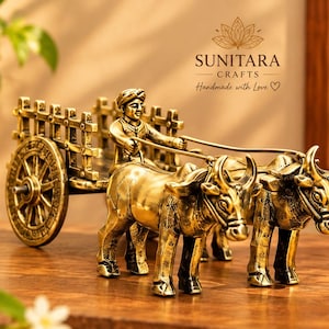 May include: A brass-colored metal figurine depicting a bullock cart pulled by two oxen with a driver seated in the cart. The piece features intricate detailing on the wheels and animals. The text SUNITARA CRAFTS Handmade with Love is visible.