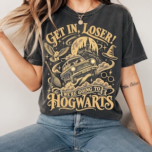 Może przedstawiać: Ciemnoszary t-shirt z krótkim rękawem ze złotym nadrukiem graficznym przedstawiającym latający samochód, zamek, sowę, miotłę, czarodziejski kapelusz i okrągłe okulary. Tekst głosi: GET IN, LOSER! WE'RE GOING TO HOGWARTS.