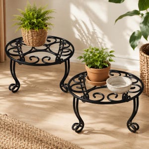 May include: Two black metal plant stands with circular tops featuring scrollwork designs and curved legs. One stand holds a green fern in a woven basket, while the other holds a green plant in a terracotta pot next to a small white bowl.