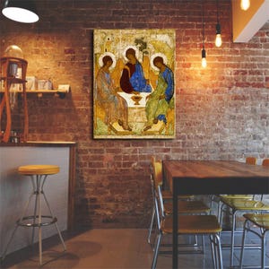 May include: A rectangular wall art print featuring a reproduction of the Holy Trinity icon. The artwork displays three figures seated around a table with a central chalice, rendered in shades of gold, blue, and red with white halos.