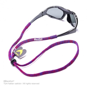 May include: Dark gray sunglasses attached to a purple and red patterned fabric strap. The strap features the white text Blackfish, a black plastic cord lock, and a small tag with a yellow logo. The eyewear has dark tinted lenses and a frame.