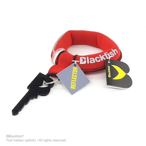 May include: A red circular fabric band with the white text Blackfish printed on it. Attached are a black plastic key-shaped accessory, a square grey tag with the yellow text REFLECTOR, and a black heart-shaped tag with a yellow logo.