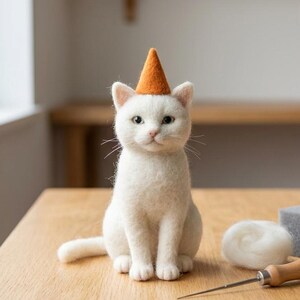 Könnte beinhalten: Eine weiße, nadelgefilzte Katzenfigur mit einem kleinen, kegelförmigen orangefarbenen Partyhut. Die Katze sitzt aufrecht mit seitlich ausgestrecktem Schwanz. Neben der Katze liegen ein kleines Knäuel weiße Wolle, eine Filznadel und ein grauer Schaumstoffblock.