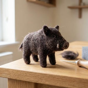 Op de afbeelding: Een donkerbruin naaldgevilte wild zwijn figuurtje met een getextureerd wollen oppervlak, kleine puntige oortjes en een korte staart. Naast het zwijn liggen een viltnaald met houten handvat, een plukje bruine wol en een grijs schuimrubberen blok.