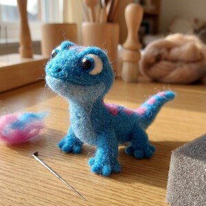 May include: A needle-felted blue lizard figurine with large white and black eyes, a light blue underbelly, and pink spots along its back and tail. A small tuft of pink and blue wool and a single metal felting needle are positioned nearby.