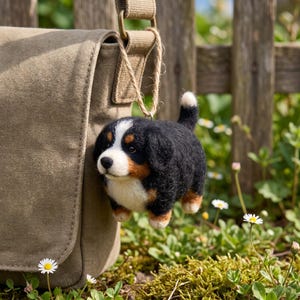 Könnte beinhalten: Ein nadelgefilzter Berner Sennenhund-Welpenanhänger mit schwarzem, weißem und braunem Fell, einer weißen Blesse im Gesicht und einer weißen Schwanzspitze, der an einer Juteschnur an einem beigen Umhängetaschenriemen aus Segeltuch hängt.
