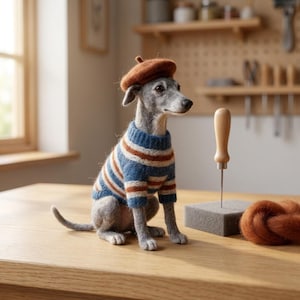 May include: A needle-felted grey Italian Greyhound figurine wearing a brown beret and a blue, white, and brown striped sweater. The figurine sits on a wooden surface next to a felting needle in a foam block and a small bundle of brown wool roving.