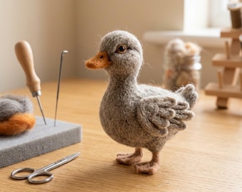 May include: A needle-felted gray duckling figurine with a textured wool finish, a small orange beak, and dark eyes. The duckling stands upright, accompanied by a gray foam pad, a wooden-handled felting needle, and a pair of small metal scissors.