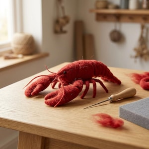 Könnte beinhalten: Eine nadelgefilzte rote Hummerfigur mit texturierter Wolloberfläche, zwei großen Vorderzangen und mehreren Beinen. Neben dem Hummer liegen eine Filznadel mit Holzgriff und ein kleines Stück grauer Schaumstoff sowie lose rote Wollfasern.