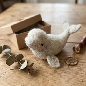 May include: A needle-felted beluga whale figurine crafted from off-white wool fibres. The whale features a rounded head, small dark eyes, side fins, and a tail fluke. It is positioned near a small cardboard box, a wax seal stamp, and a sprig of eucalyptus.
