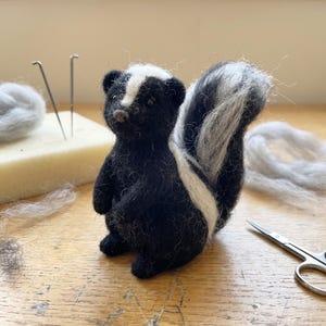May include: A needle-felted skunk figurine crafted from black and white wool fibres. The small animal features a prominent white stripe running from its forehead down its back, ending in a fluffy, upright tail with white and black textured fur.