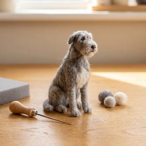May include: A needle-felted gray and white dog figurine sitting upright. The dog has a textured, woolly coat with a lighter patch on its chest. Beside the figurine are three small wool balls and a wooden-handled metal felting needle tool.