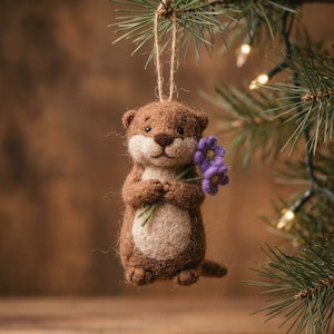 May include: A needle-felted brown otter ornament with a cream-colored belly, holding a small bunch of purple flowers. The otter has dark eyes and a brown nose, and is suspended by a thin jute twine loop for hanging on a tree branch.