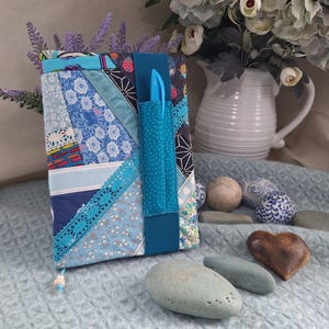 May include: A fabric-covered journal featuring a patchwork design in various shades of blue, white, and teal. The cover includes a vertical teal elastic band with an integrated pen holder containing a blue pen and a decorative lace trim accent.