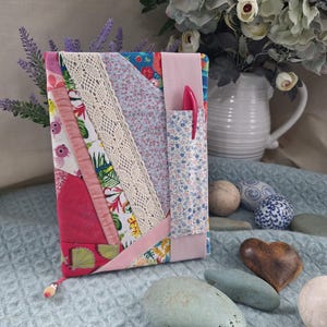 May include: A fabric-covered journal featuring a patchwork design of floral and solid pink panels, accented with a strip of cream-colored lace and a vertical light pink fabric band. A small floral fabric pocket holds a bright pink pen on the front.