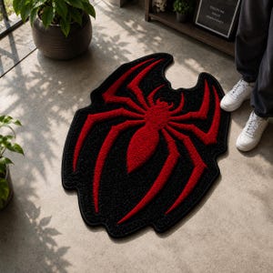 May include: A tufted area rug featuring a bold, red spider design centred on a black background. The rug has an irregular, organic shape with a thick black border, showcasing a textured, plush pile surface that contrasts the vibrant red spider.