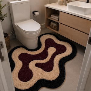 May include: An irregular, wavy-shaped bathroom rug featuring a layered, abstract pattern. The rug has a deep burgundy center, surrounded by a thick band of cream-colored textured material, and finished with a solid black border edge.