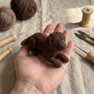 Könnte beinhalten: Eine kleine, handgefertigte Welpenfigur aus brauner Nadelfilzwolle mit strukturierter Oberfläche, dunklen Augen und einer weichen, abgerundeten Schnauze. Der Miniaturhund ist in liegender Position dargestellt und zeigt detaillierte Handwerkskunst.