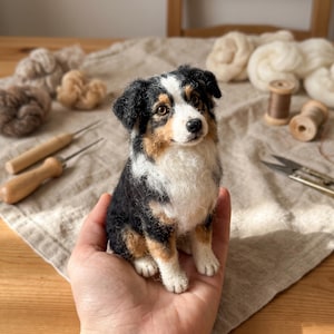 Puede incluir: Una figura de perro Pastor Australiano hecha con lana fieltrada con aguja, con un pelaje tricolor en negro, blanco y canela. El perro en miniatura tiene una mancha blanca en la cara, ojos marrón oscuro y nariz negra, con acabado texturizado.