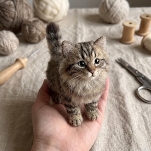 Peut inclure: Une petite figurine de chaton en laine feutrée avec un pelage tigré brun et noir, des oreilles pointues et des yeux sombres. Le chat miniature a un aspect texturé et duveteux avec une queue courte et droite et des traits détaillés.