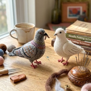 Könnte beinhalten: Zwei nadelgefilzte Vogelfiguren, eine graue mit irisierenden Halsdetails und einem kleinen Wurm im Schnabel sowie eine reinweiße. Sie stehen auf einer Holzoberfläche neben einer Keramiktasse, Büchern und verschiedenen Bastelwerkzeugen.