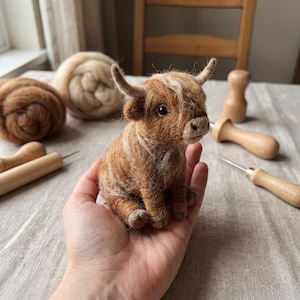 Könnte beinhalten: Eine kleine, nadelgefilzte Highland-Rinderfigur mit zotteligem, hell- und dunkelbraunem Wollvlies, kleinen geschwungenen Hörnern und dunklen Augen. Das Miniaturtier ist mit texturiertem, realistisch wirkendem Fell detailliert gestaltet.