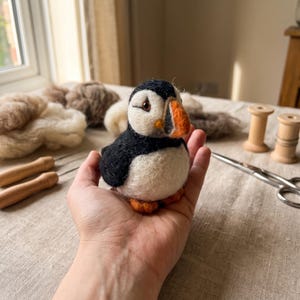 May include: A needle-felted puffin figurine featuring a black and white body, a bright orange beak with grey accents, and small orange feet. The bird has a rounded, soft texture and is crafted from wool fibres, showcasing detailed facial features.