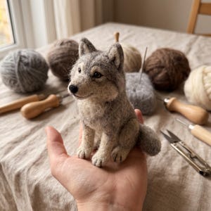 Peut inclure: Une figurine de loup en laine feutrée à l'aiguille dans des tons gris, blancs et beige. La petite sculpture présente des oreilles pointues, un nez noir et des yeux sombres, représentant un loup assis avec un pelage texturé et une queue touffue.