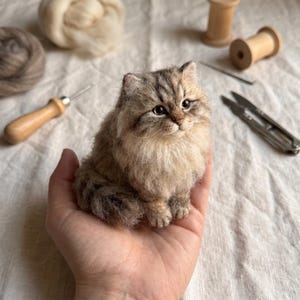 Peut inclure: Une petite figurine de chat feutrée à l'aiguille, fabriquée à la main, avec une fourrure longue et duveteuse de couleur brun clair et crème, des marques tabby brun foncé sur la tête et des yeux sombres réalistes, sculptée en position assise.