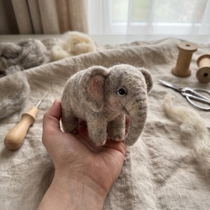 Könnte beinhalten: Eine nadelgefilzte Elefantenfigur aus melierten grauen und weißen Wollfasern. Die kleine Skulptur verfügt über große Ohren mit zarten rosa Innendetails, einen Rüssel und dunkle Perlenaugen, die eine texturierte, flauschige Oberfläche aufweisen.