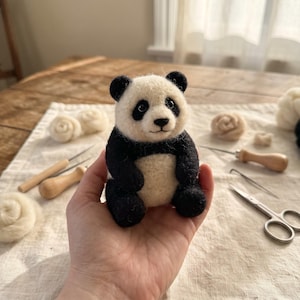 Könnte beinhalten: Eine kleine, handgefertigte, nadelgefilzte Panda-Figur mit cremefarbenem Gesicht und Bauch, schwarzen Ohren, schwarzen Augenpartien und schwarzen Gliedmaßen. Der Panda hat einen runden Kopf, eine kleine schwarze Nase und einen gestickten Mund.