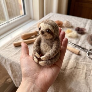 May include: A small, needle-felted sloth figurine crafted from textured, light and dark brown wool fibers. The sloth features a rounded head, dark bead eyes, a small nose, and long, curved arms with light-colored claws, sitting in a seated position.