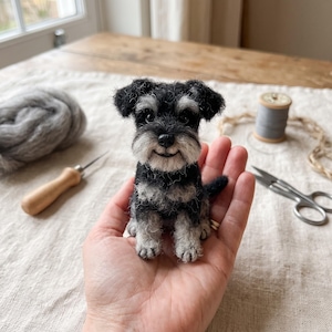 Op de afbeelding: Een naaldgevilte miniatuur Schnauzer hondje met zwart en zilvergrijs bont, voorzien van hangende oren, een textuurrijke vacht en een kleine zwarte neus, met realistische details vervaardigd om op een zittende puppy te lijken.