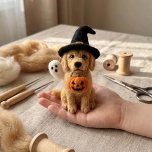 Puede incluir: Una figura de cachorro dorado hecha con fieltro de aguja que lleva un sombrero de bruja negro, sostiene una pequeña calabaza naranja en la boca y tiene un pequeño fantasma blanco unido al costado, creada con fibras de lana texturizadas.