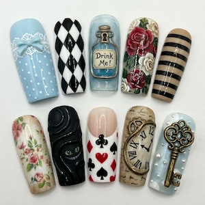 K&ouml;nnte beinhalten: Ein Set aus zehn k&uuml;nstlichen Fingern&auml;geln mit verschiedenen Designs, darunter hellblaue Punkte mit einer Schleife, schwarze und wei&szlig;e Rauten, eine blaue Flasche mit der Aufschrift Drink Me!, rote und wei&szlig;e Rosen, schwarze Streifen, ein Katzengesicht, Spielkartensymbole, eine Uhr und ein Schl&uuml;ssel.