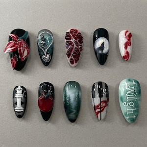May include: A set of ten almond-shaped press-on nails featuring diverse designs including a red flower, a heart locket, pomegranate seeds, a wolf howling at the moon, red swirls, a white chess piece, a red apple, a forest, piano keys, and the word Twilight.