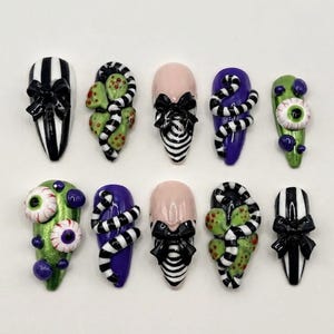 May include: A set of ten press-on nails featuring varied designs including black and white vertical stripes with black bows, green textured surfaces with eyeballs, purple bases with black and white striped snakes, and nude bases with black bows.