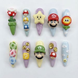 May include: A set of ten press-on nails featuring 3D resin charms inspired by video game characters and items, including Mario, Luigi, Piranha Plant, stars, mushrooms, coins, question blocks, and clouds in vibrant colors like red, green, and yellow.