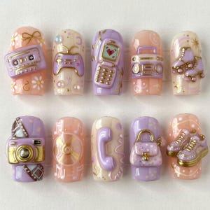 May include: A set of ten press-on nails featuring 3D relief designs in pastel purple, pink, and cream. The motifs include a cassette tape, game controller, flip phone, boombox, roller skates, camera, CD, telephone handset, handbag, and sneakers.